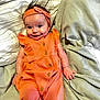 baby, bed, blanket, buttoned_clothing, child, cozy, cuteness, hand, happy, headband, indoors, infant, lying_down, orange_outfit, person, portrait, skin, smile, soft_fabric, young