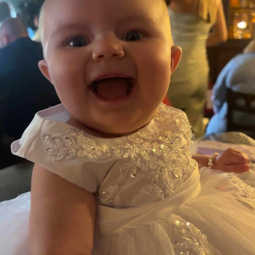 Saoirse is registered to the contest to win money with this photo: baby, child, smiling, dress, white_dress, indoor, party, celebration, person, happy, face, portrait, people, event, blurred_background, infant, cute, toddler, formal_wear, fun