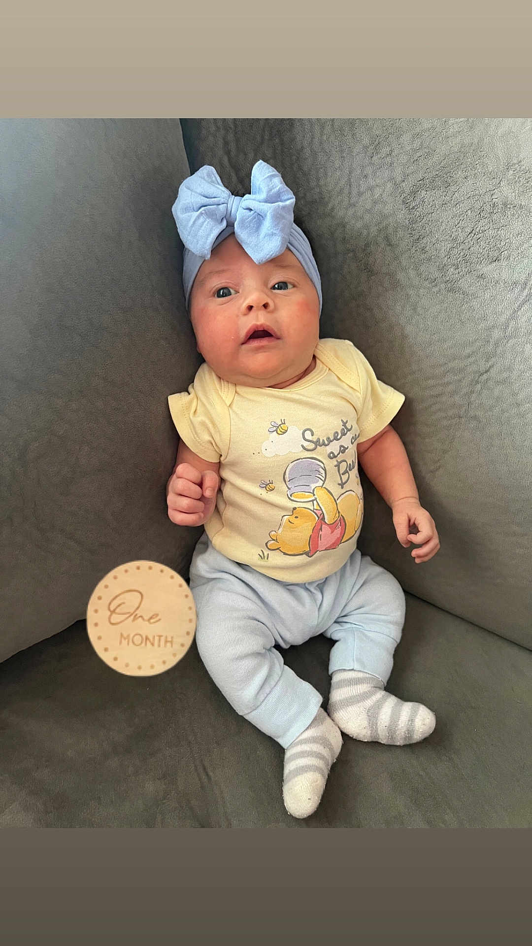 Avery is registered to the contest to win money with this photo: baby, infant, one_month, headband, bow, yellow_onesie, winnie_the_pooh, couch, gray_couch, striped_socks, wooden_milestone, milestone_marker, cute, portrait, seated, indoors, child, smiling, soft_lighting, cozy