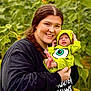 woman, baby, child, sunflower, flower, green_clothing, outdoor, nature, smile, holding, portrait, plants, field, happy, person, infant, headwear, bright_colors, grass, casual_clothing