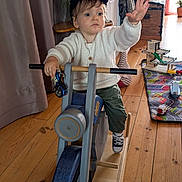 Charles participe au concours pour gagner de l'argent avec cette photo : toddler, child, indoor, wooden_toy, rocking_motorcycle, toy_bike, white_sweater, cap, wooden_floor, playmat, toys, reaching_hand, determined, casual_clothing, playtime, furniture, plant, curtain, cabinet, home