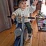 toddler, child, indoor, wooden_toy, rocking_motorcycle, toy_bike, white_sweater, cap, wooden_floor, playmat, toys, reaching_hand, determined, casual_clothing, playtime, furniture, plant, curtain, cabinet, home