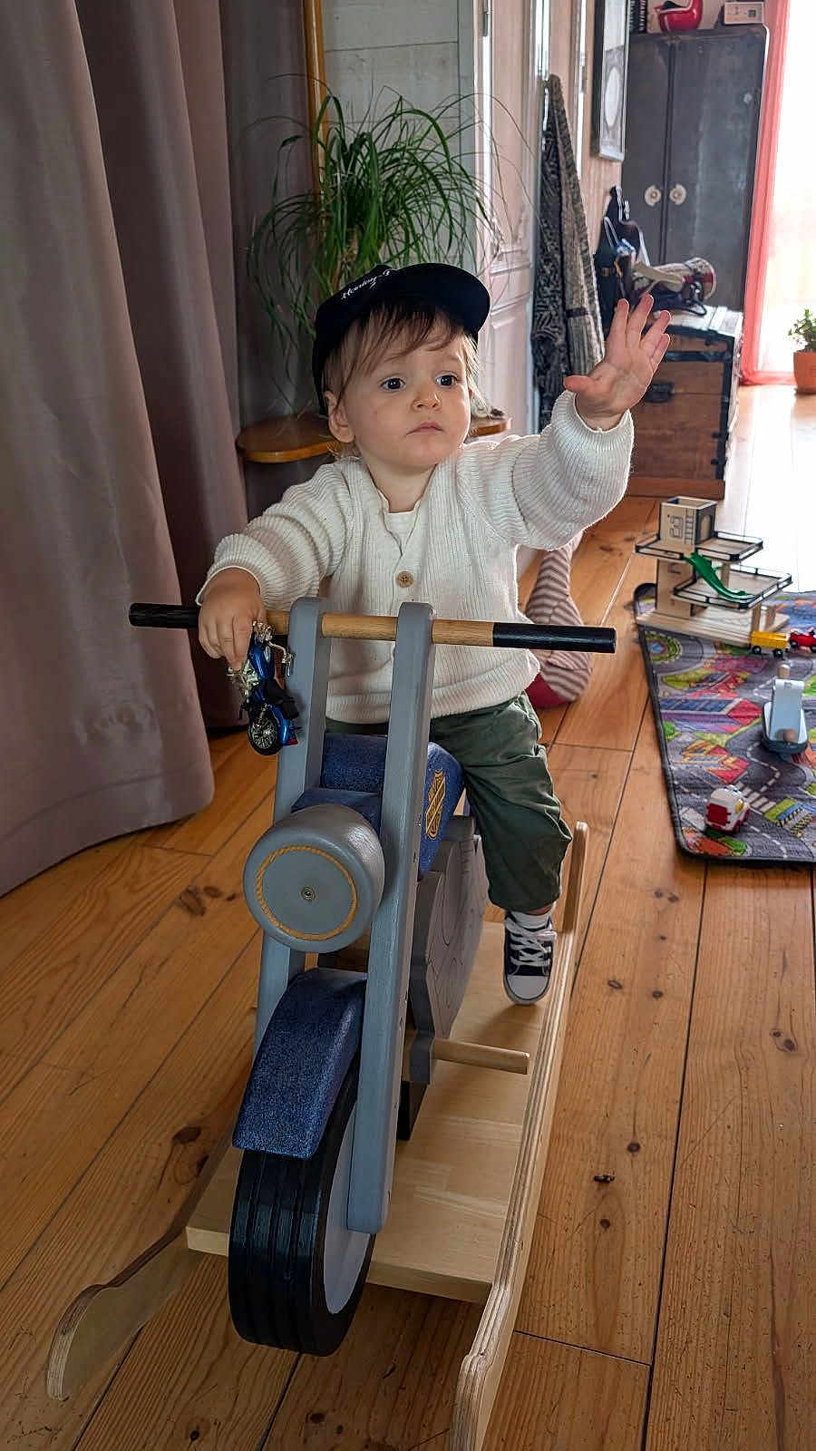 Charles participe au concours pour gagner de l'argent avec cette photo : toddler, child, indoor, wooden_toy, rocking_motorcycle, toy_bike, white_sweater, cap, wooden_floor, playmat, toys, reaching_hand, determined, casual_clothing, playtime, furniture, plant, curtain, cabinet, home