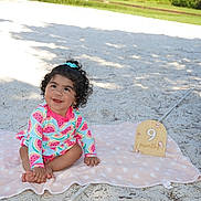 Sophia is registered to the contest to win money with this photo: baby, smiling, sitting, blanket, sand, outdoor, watermelon_pattern, curly_hair, child, cute, happy, nature, greenery, park, sunlight, wooden_sign, nine_months, footwear_free, playful, summer