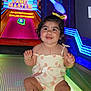 toddler, child, smiling, bowling_lane, arcade, colorful, glowing, yellow_bow, curly_hair, happy, indoor, playful, bright_colors, signage, toy, footwear, cute, entertainment, small_child, sitting