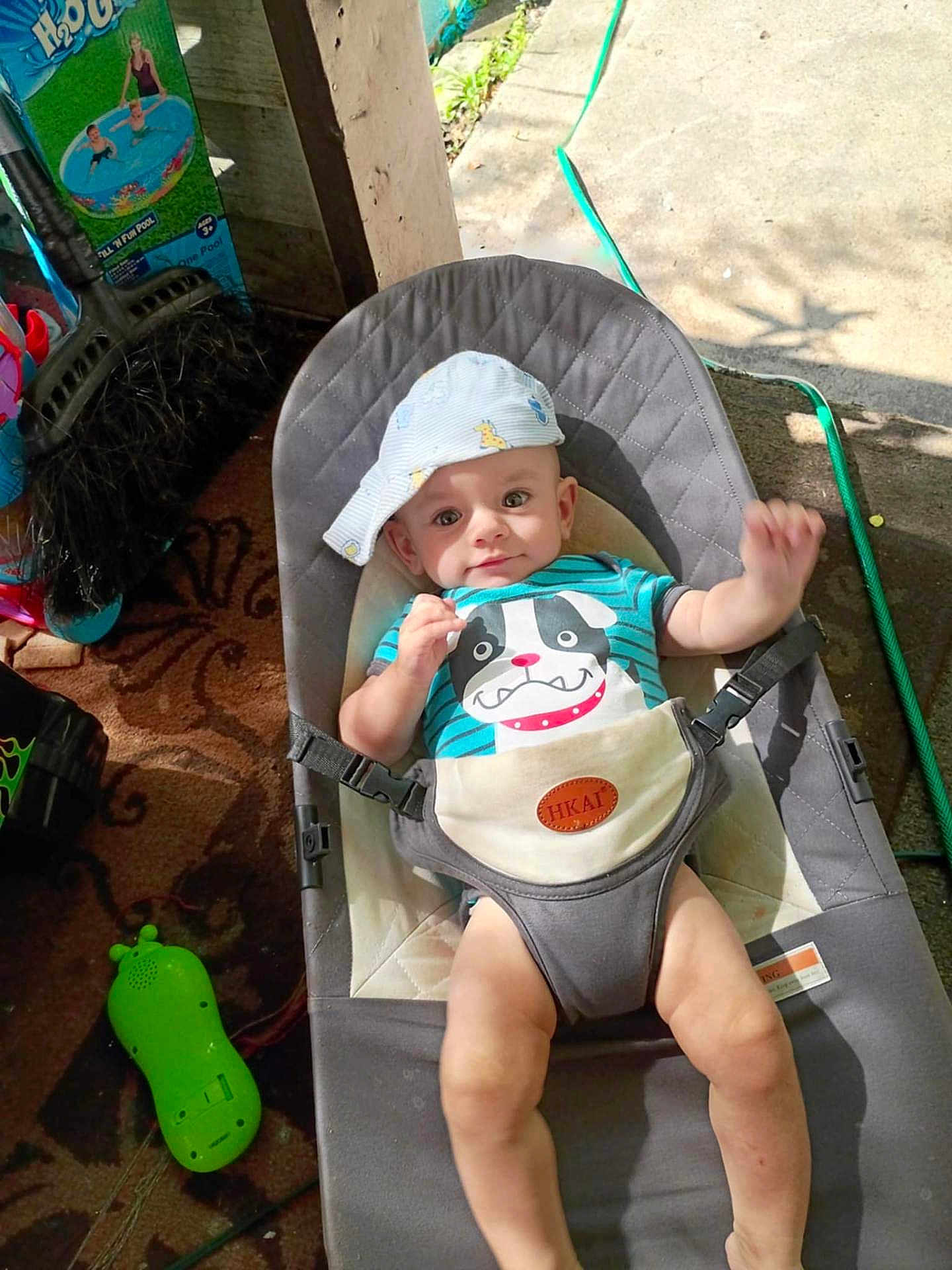 Levi joined the competition — help win amazing prizes! baby, child, infant, baby_bouncer, hat, clothing, shirt, toy, outdoor, sunlight, shadow, hose, floor, carpet, person, smiling, face, legs, hand, seat