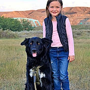 Ruger is registered to the contest to win money with this photo: child, dog, black_dog, grass, field, outdoor, smile, happy, girl, casual_clothing, boots, vest, jeans, leash, nature, bridge, hills, sky, sunset_light, pet