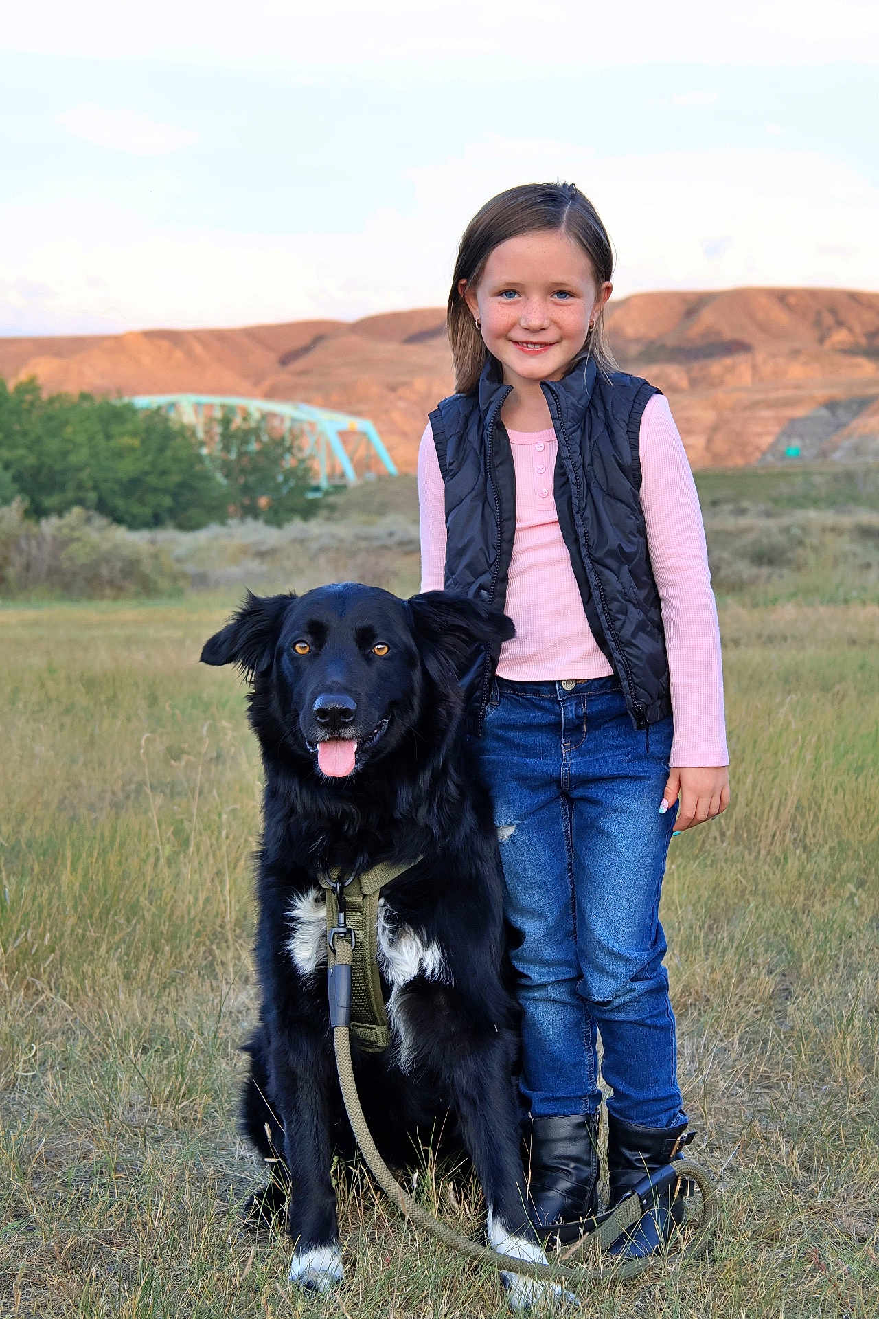 Ruger is registered to the contest to win money with this photo: child, dog, black_dog, grass, field, outdoor, smile, happy, girl, casual_clothing, boots, vest, jeans, leash, nature, bridge, hills, sky, sunset_light, pet
