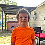 child, boy, curly_hair, orange_shirt, backyard, patio, shed, garden_furniture, red_cushion, outdoor, smile, bracelet, daylight, casual, standing, concrete_floor, window_air_conditioner, wooden_fence, trees, relaxed