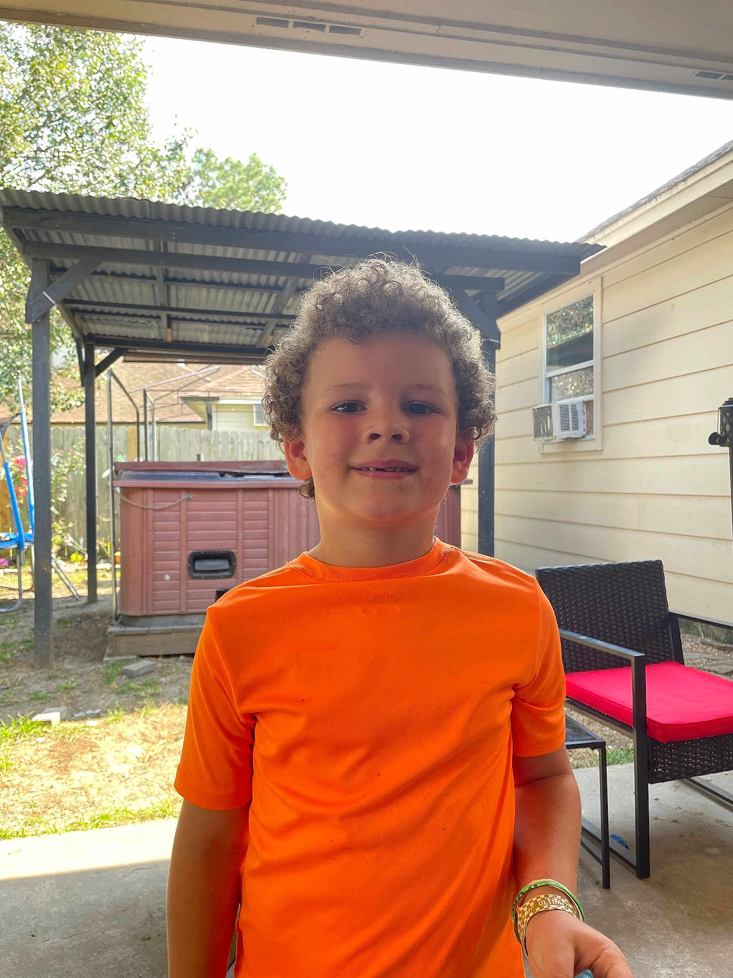Daniel joined the competition — help win amazing prizes! child, boy, curly_hair, orange_shirt, backyard, patio, shed, garden_furniture, red_cushion, outdoor, smile, bracelet, daylight, casual, standing, concrete_floor, window_air_conditioner, wooden_fence, trees, relaxed
