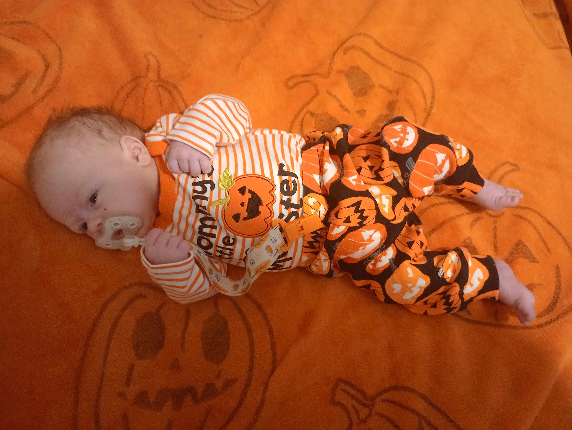 Giovanni is registered to the contest to win money with this photo: baby, infant, pacifier, orange_blanket, pumpkin_pattern, halloween_theme, clothing, sleepwear, face, head, hands, feet, lying_down, cute, child, young, indoor, blanket, pattern, festive