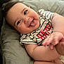 arm, baby, casual_clothing, child, chubby_cheeks, closeup, cute, face, happy, indoors, infant, lying_down, playful, portrait, red_text, skin, smiling, soft_surface, striped_shirt, tongue_out