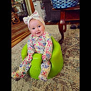 Sophia is registered to the contest to win money with this photo: baby, child, smiling, headband, onesie, green_baby_seat, indoor, carpet, wood_floor, table, basket, furniture, living_room, happy, cute, sitting, portrait, colorful_clothing, person, home