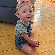 Macie is registered to the contest to win money with this photo: toddler, child, pink_bow, overalls, orange_ball, wooden_floor, indoor, living_room, couch, smile, sitting, curious, light, small_person, casual_clothing, short_sleeves, floor, cute, young_child, home