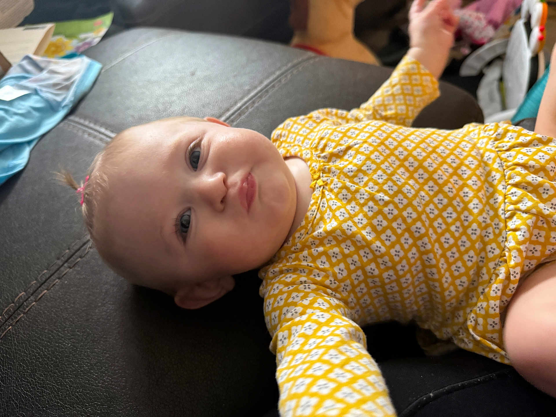 Macie is registered to the contest to win money with this photo: baby, infant, child, yellow_clothing, patterned_outfit, lying_down, leather_surface, funny_face, pursed_lips, cute, indoor, person, head, arm, skin, eyes, hair, expression, closeup, casual
