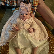 Skyla joined the competition — help win amazing prizes! adult_hand, baby, blanket, bow, clothing, cozy, cute, dress, fabric, floor, headband, indoor, infant, legs, lying_down, person, plaid_shirt, skin, soft_texture, wood_floor