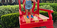 Andre is registered to the contest to win money with this photo: toddler, child, outdoor, red_chair, wooden_chair, greenery, shoes, denim_overalls, sky, clouds, tree, plants, side_view, curious, standing, daylight, nature, playful, young_child, casual_clothing