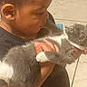 child, cat, gray_cat, white_paws, holding, indoor, tiled_floor, gentle, affection, curiosity, young_child, pet, animal, human_hand, close_up, face, interaction, soft_lighting, caring, bond