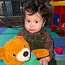 toddler, child, curly_hair, teddy_bear, toy, play_mat, indoor, baby_gate, colorful, cute, clothing, overalls, expression, face, person, playroom, smile, young_child, plush_toy, seated