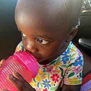 Marsai is registered to the contest to win money with this photo: toddler, child, sippy_cup, drinking, colorful_clothing, floral_pattern, cute, indoors, portrait, baby, hand, face, skin, hair, curious, bright, natural_light, closeup, young, casual