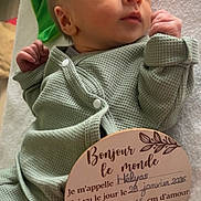Hélyas participe au concours pour gagner de l'argent avec cette photo : baby, newborn, infant, face, eyes_open, looking_up, onesie, green_clothing, blanket, wooden_sign, hand, soft_texture, portrait, indoor, cozy, changing_table_surface, handwritten_text, adorable, small_hands, cute