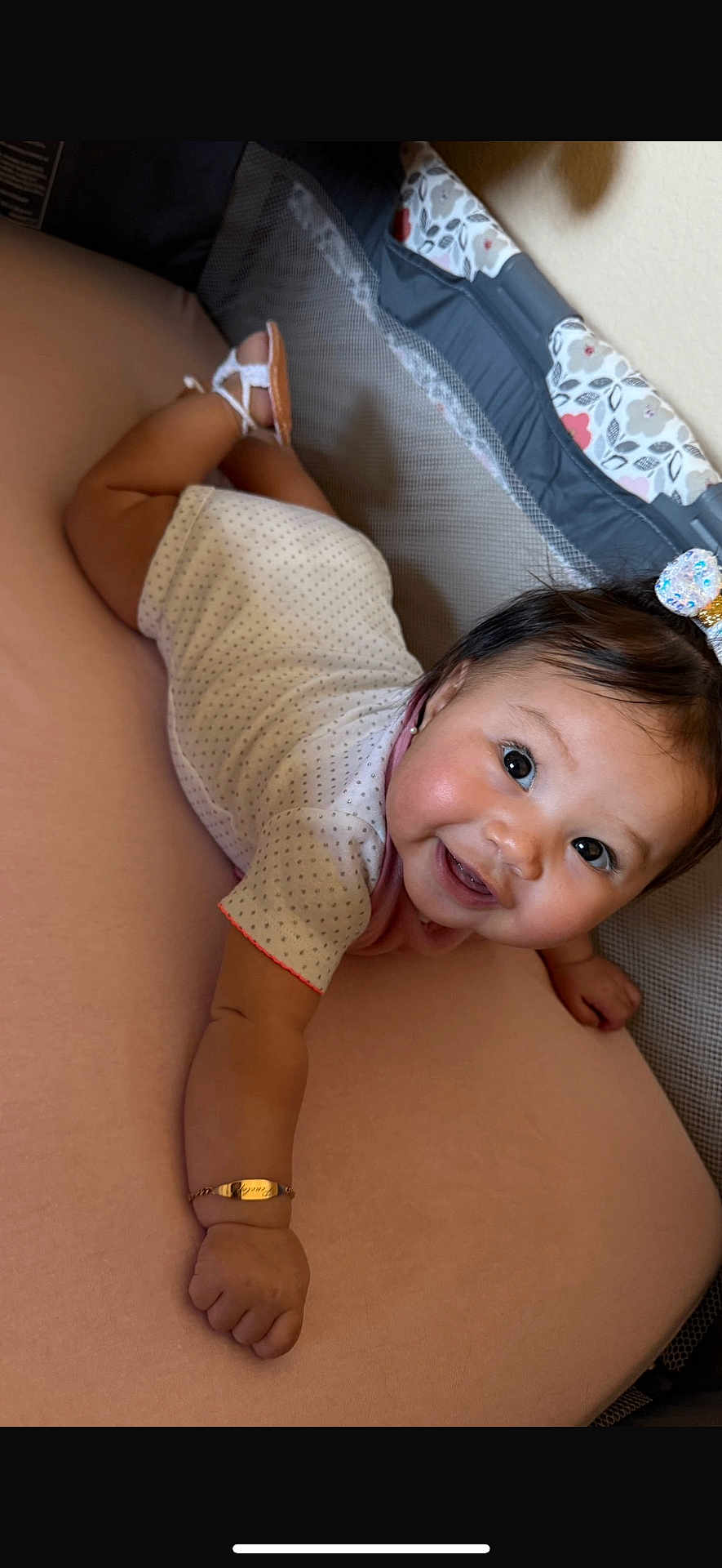 Penelope is registered to the contest to win money with this photo: baby, smiling, face, arm, hand, bracelet, hair_bow, white_clothing, polka_dots, pink_mattress, lying_down, indoors, happy, cute, child, infant, skin, person, closeup, portrait