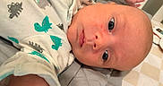 Theodore is registered to the contest to win money with this photo: baby, infant, face, onesie, dinosaur_pattern, blue_eyes, lying_down, cute, newborn, child, skin, head, clothing, bed, blanket, indoors, portrait, expression, closeup, person