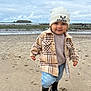beach, beanie, boots, child, cloudy_sky, coast, curly_hair, footwear, jeans, nature, ocean, outdoor, person, plaid_jacket, portrait, sand, smile, sweater, toddler, water