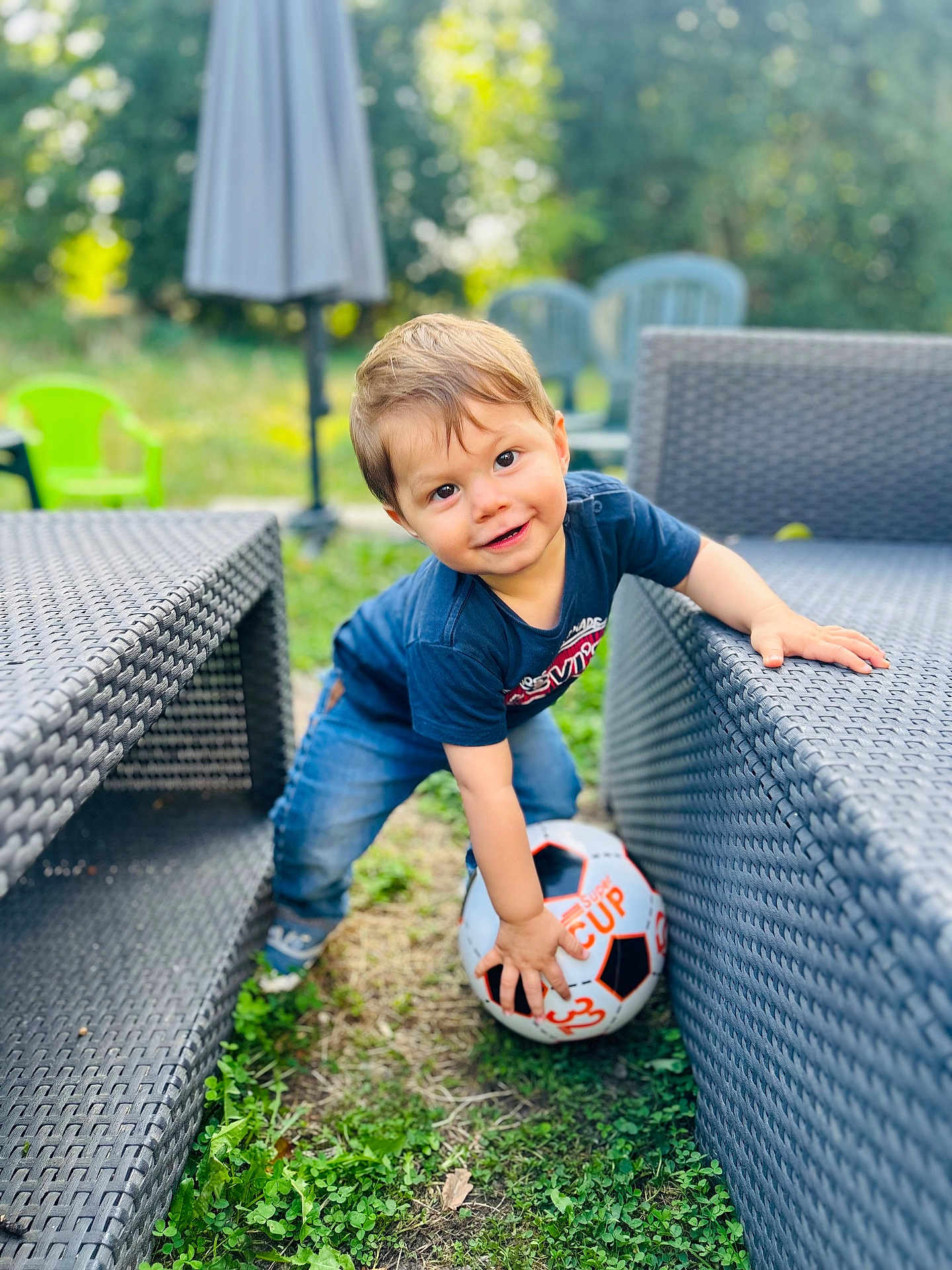 Raphaël participe au concours pour gagner de l'argent avec cette photo : toddler, child, soccer_ball, outdoor, grass, patio_furniture, woven_chair, blue_shirt, jeans, smiling, playing, greenery, umbrella, plastic_chair, daylight, happy, cute, person, young_child, fun