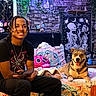 bed, blanket, braided_hair, casual, colorful, dog, furniture, happy, indoor, patterned_bedding, person, pet, pillow, portrait, room, smiling, string_lights, tapestry, tattoo, wall_decor