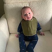 Rowan is registered to the contest to win money with this photo: baby, infant, child, sitting, armchair, bib, onesie, sports_pattern, blue_clothing, green_bib, cozy, indoor, soft_texture, curious_expression, wide_eyes, comfortable, portrait, young_child, cute, home