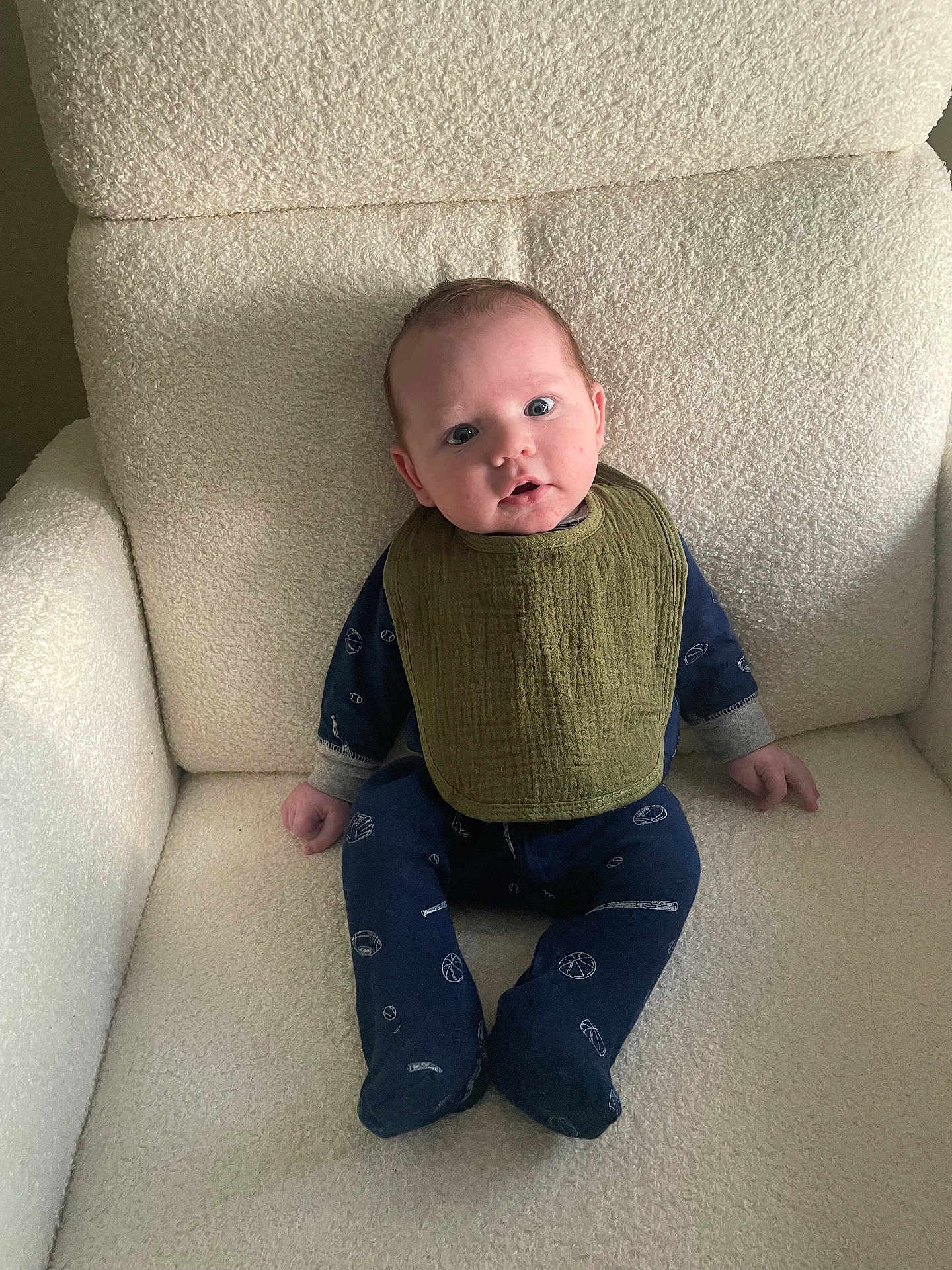 Rowan is registered to the contest to win money with this photo: baby, infant, child, sitting, armchair, bib, onesie, sports_pattern, blue_clothing, green_bib, cozy, indoor, soft_texture, curious_expression, wide_eyes, comfortable, portrait, young_child, cute, home