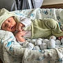 baby, sleeping, green_clothing, hat, basket, blanket, golf_balls, indoor, cozy, infant, soft_texture, cute, resting, child, person, closeup, home, floor, relaxing, peaceful