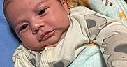 Andres is registered to the contest to win money with this photo: baby, infant, child, portrait, close_up, face, eyes_open, lying_down, blanket, onesie, clothing, blue_cushion, plush_fabric, indoor, home, cozy, relaxed, head, skin, photography