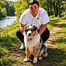 Baïko a rejoint le concours — aidez-le/la à gagner de superbes lots ! man, person, dog, australian_shepherd, pet, outdoor, riverside, river, forest, trees, nature, walking_trail, grass, sunlight, portrait, crouching, white_shirt, black_pants, collar, leash