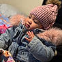 baby, beanie, bodypart, bonnet, cap, clothing, coat, couch, face, finger, hand, hat, head, hoodie, jacket, knitwear, pants, person, sweater, sweatshirt
