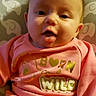 baby, infant, child, pink_sweatshirt, text_on_clothing, born_wild, leopard_print, cushion, elephant_pattern, tongue_out, happy, cute, face, portrait, sitting, indoors, person, clothing, head, smile