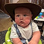 baby, child, cowboy_hat, baby_seat, indoor, curtains, toy, table, face, blue_eyes, chubby_cheeks, white_tshirt, denim_overalls, hand, home_interior, framed_photo, curious_expression, furniture, young_child, seat