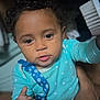 toddler, child, baby, curly_hair, blue_onesie, pacifier_clip, hand, face, eyes, indoor, person, closeup, soft_lighting, expression, holding, reaching, brush, background_blur, skin, cute
