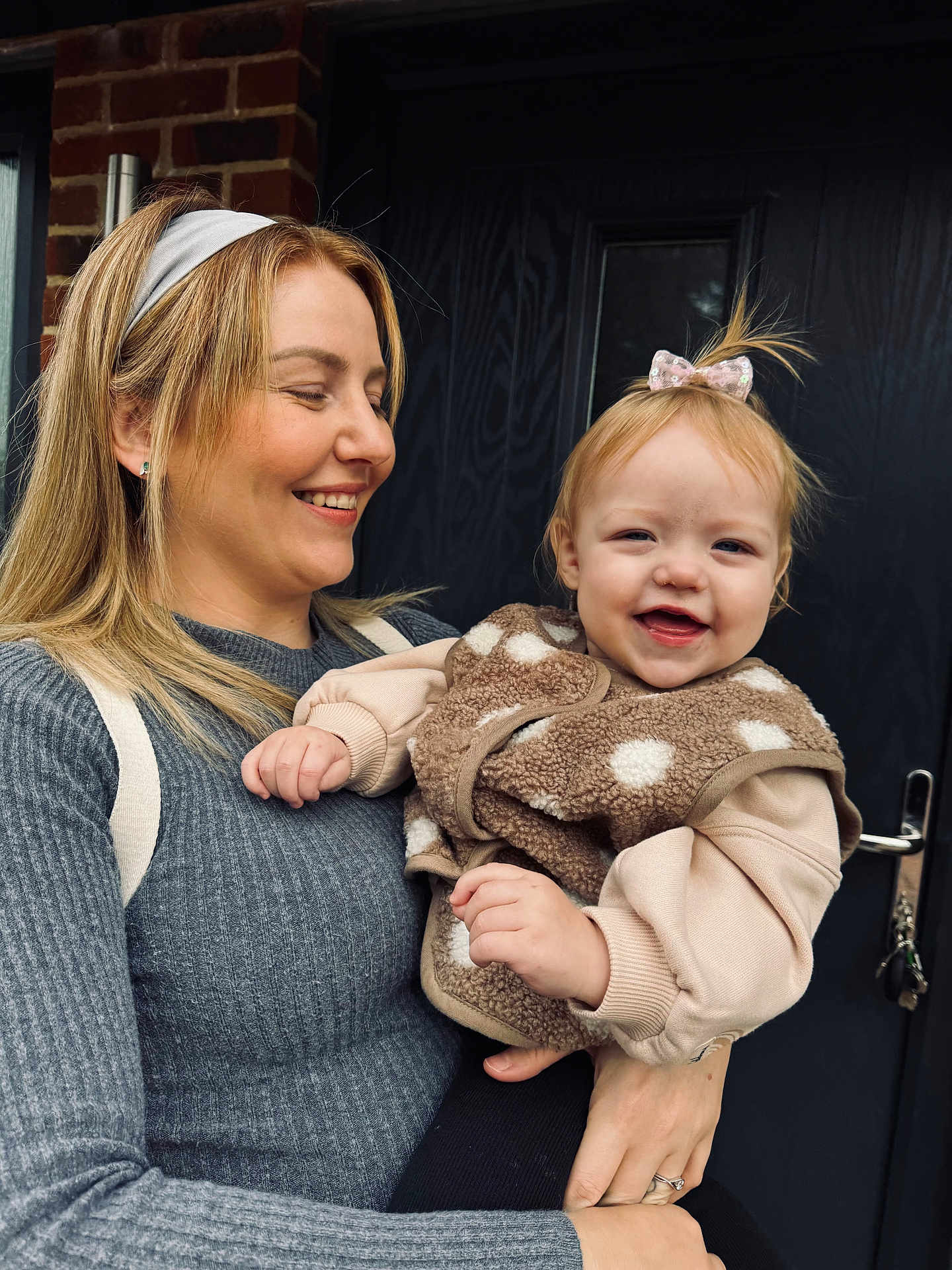 Willow is registered to the contest to win money with this photo: woman, toddler, child, smile, holding, hair_bow, gray_sweater, door, brick_wall, happy, portrait, outdoor, person, clothing, baby, casual, family, cute, headband, warm_clothing