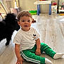 black_cat, cat, child, clothing, cute, floor, furniture, green_pants, indoor, mickey_mouse, person, pet, sitting, smile, socks, toddler, toy, white_tshirt, window, wooden_floor
