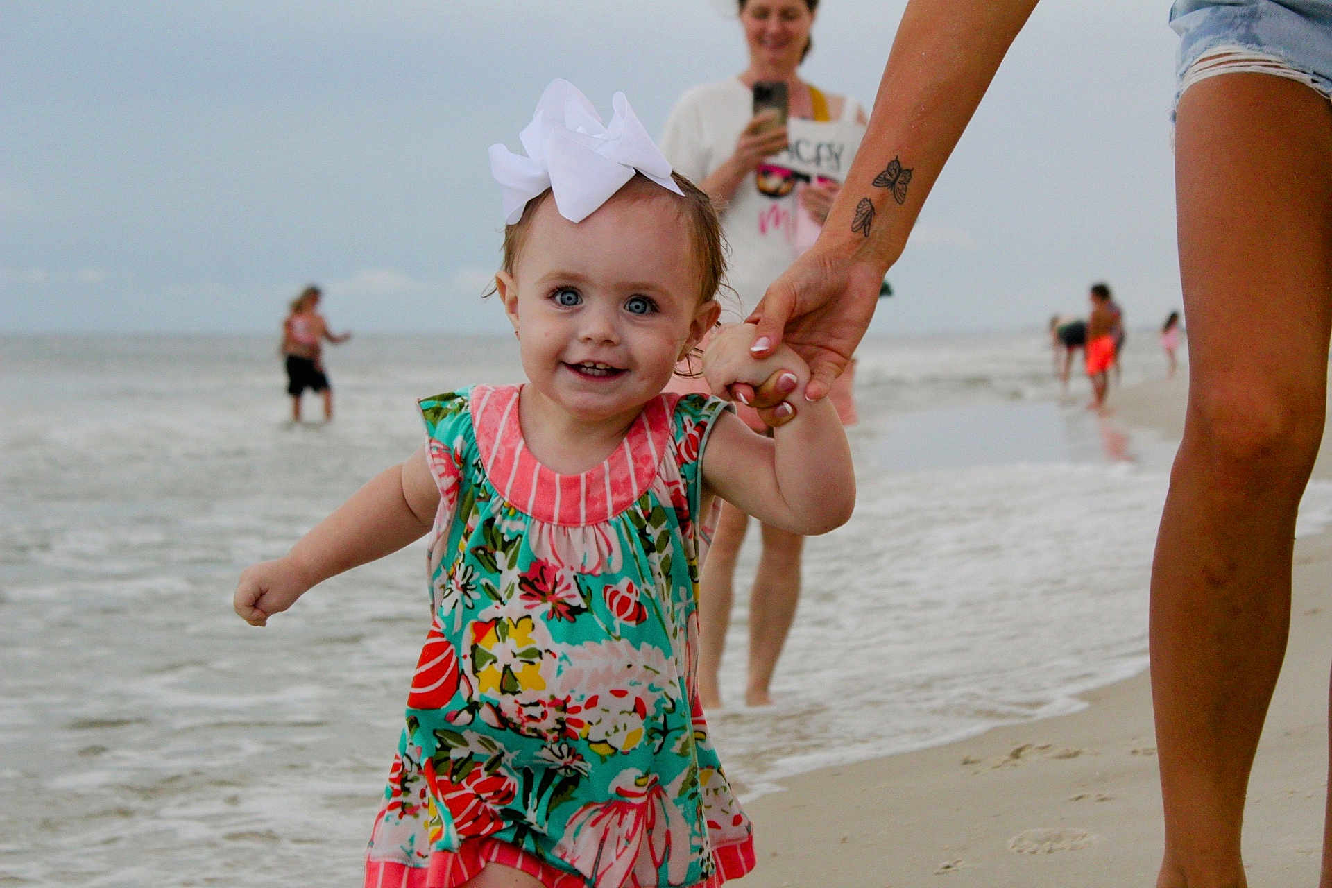 Berkleigh Grace joined the competition — help win amazing prizes! toddler, child, beach, water, ocean, sand, hand_holding, bow, colorful_dress, smiling, happy, adult, legs, tattoo, people, background, cloudy_sky, shoreline, family, vacation