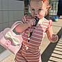 toddler, child, pink_purse, striped_clothing, outdoor, sunlight, shadows, car_key, hair_bow, footwear, curious_expression, standing, young_child, casual_clothing, playful, person, daylight, concrete_floor, decor, accessories