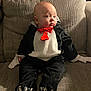 baby, child, penguin_costume, red_bow_tie, couch, beige, texture, seated, indoor, costume, cute, expression, curious, clothing, footwear, hands, head, face, person, soft_lighting
