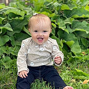 Cora is registered to the contest to win money with this photo: baby, child, infant, smiling, laughing, portrait, outdoor, garden, grass, plants, leaves, wooden_fence, barefoot, hair_bow, floral_shirt, long_sleeves, dark_pants, sitting, happy, nature