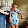toddler, child, person, baby, blue_outfit, standing, indoor, adult_hand, jeans, long_sleeve_shirt, furniture, chair, couch, expression, face, cute, home, smiling, holding, portrait
