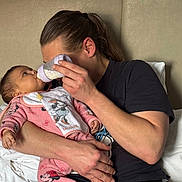 Aaliyah a rejoint le concours — aidez-le/la à gagner de superbes lots ! adult, baby, baby_bottle, bed, bib, cuddling, feeding, gaze, hair_tie, hand, headboard, holding, indoor, infant, loving, man, minnie_mouse, pajamas, pillow, portrait