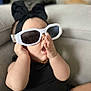 baby, infant, sunglasses, headband_bow, black_outfit, arm, hand, couch, living_room, portrait, close_up, cute, accessories, eyewear, open_mouth, soft_focus, seat, necklace, fashion, indoor