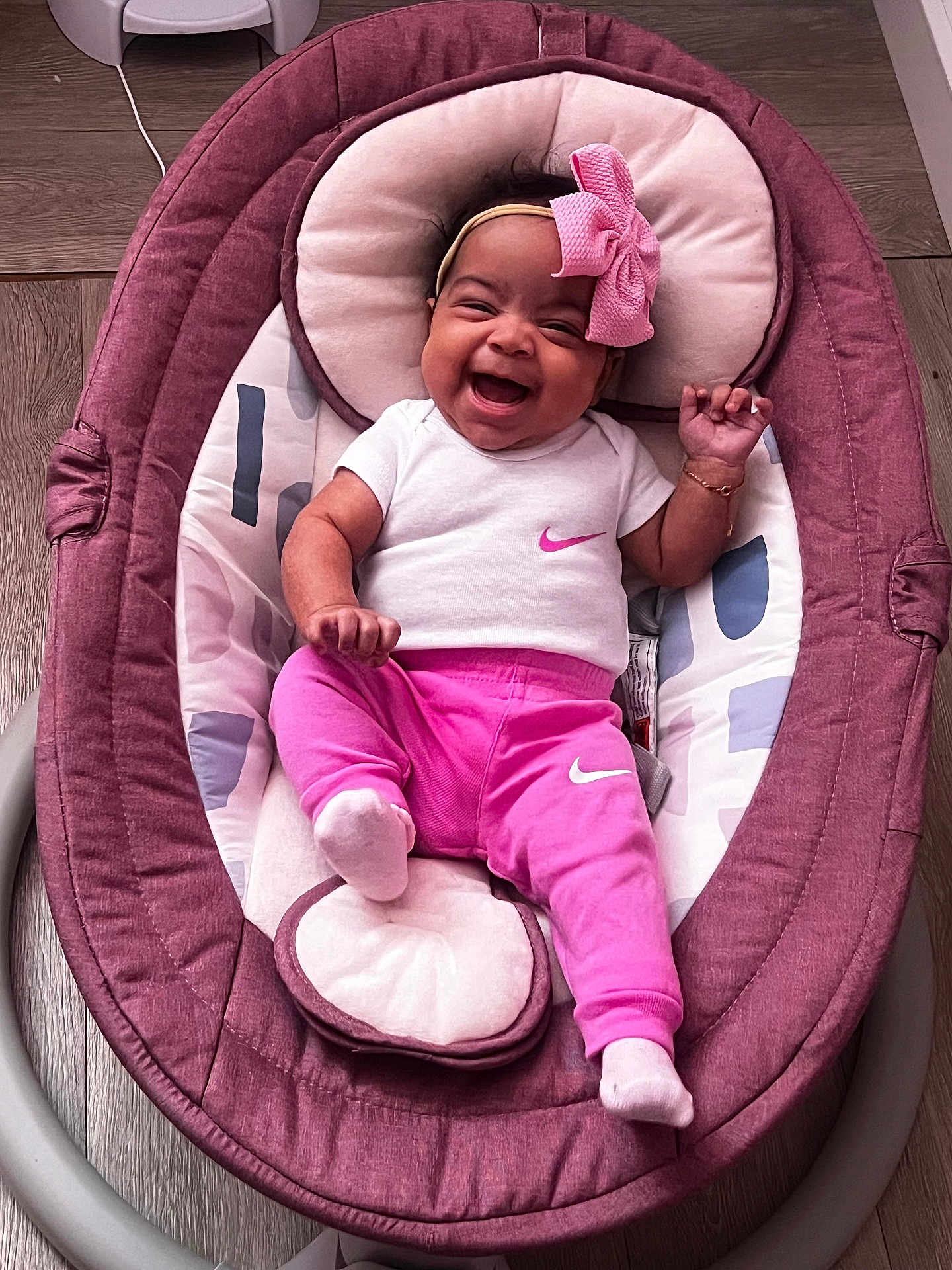 Emma is registered to the contest to win money with this photo: baby, infant, smiling, happy, pink_clothing, white_shirt, headband, bow, baby_rocker, indoor, wooden_floor, footwear, sock, child, person, cute, portrait, relaxing, apparel, joy