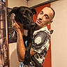 Princesse participe au concours pour gagner de l'argent avec cette photo : man, dog, indoor, holding, black_dog, short_hair, tattoo, tshirt, wall_art, door, portrait, pet, animal, casual, human, expression, arm, furniture, home, room